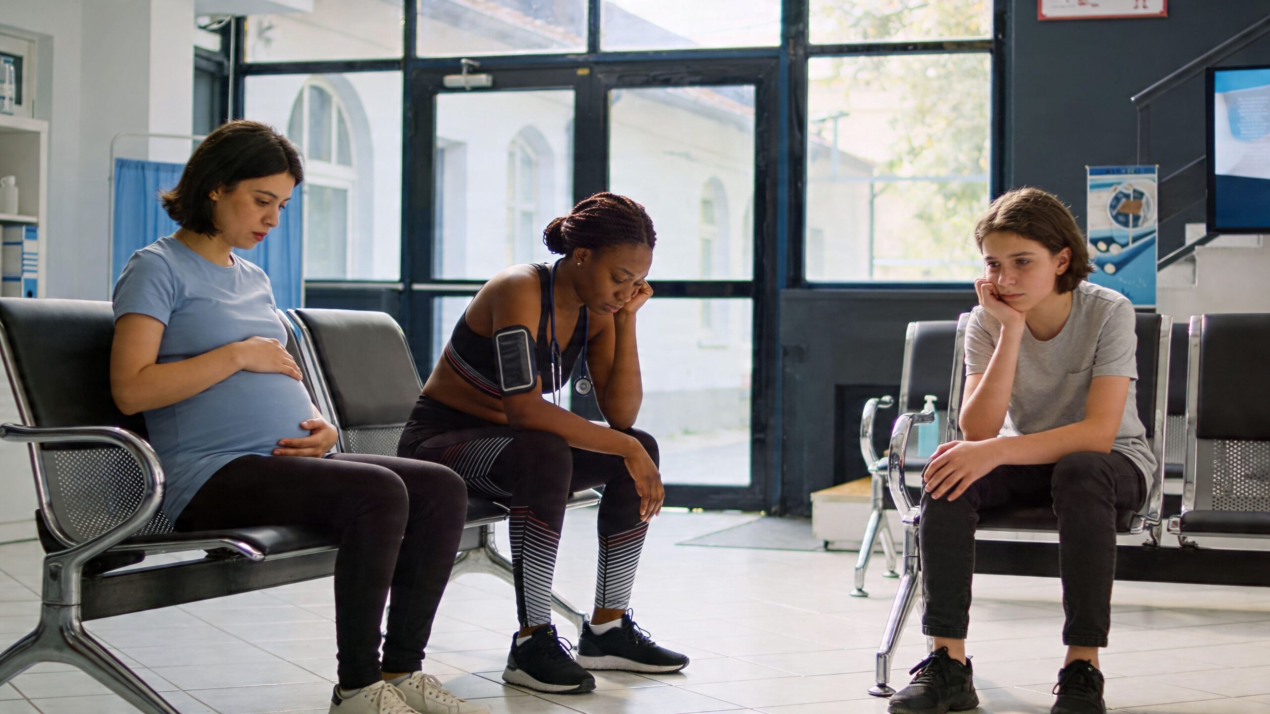 A pregnant woman, a woman in activewear, and a boy sitting dejectedly in a quiet medical office waiting room.