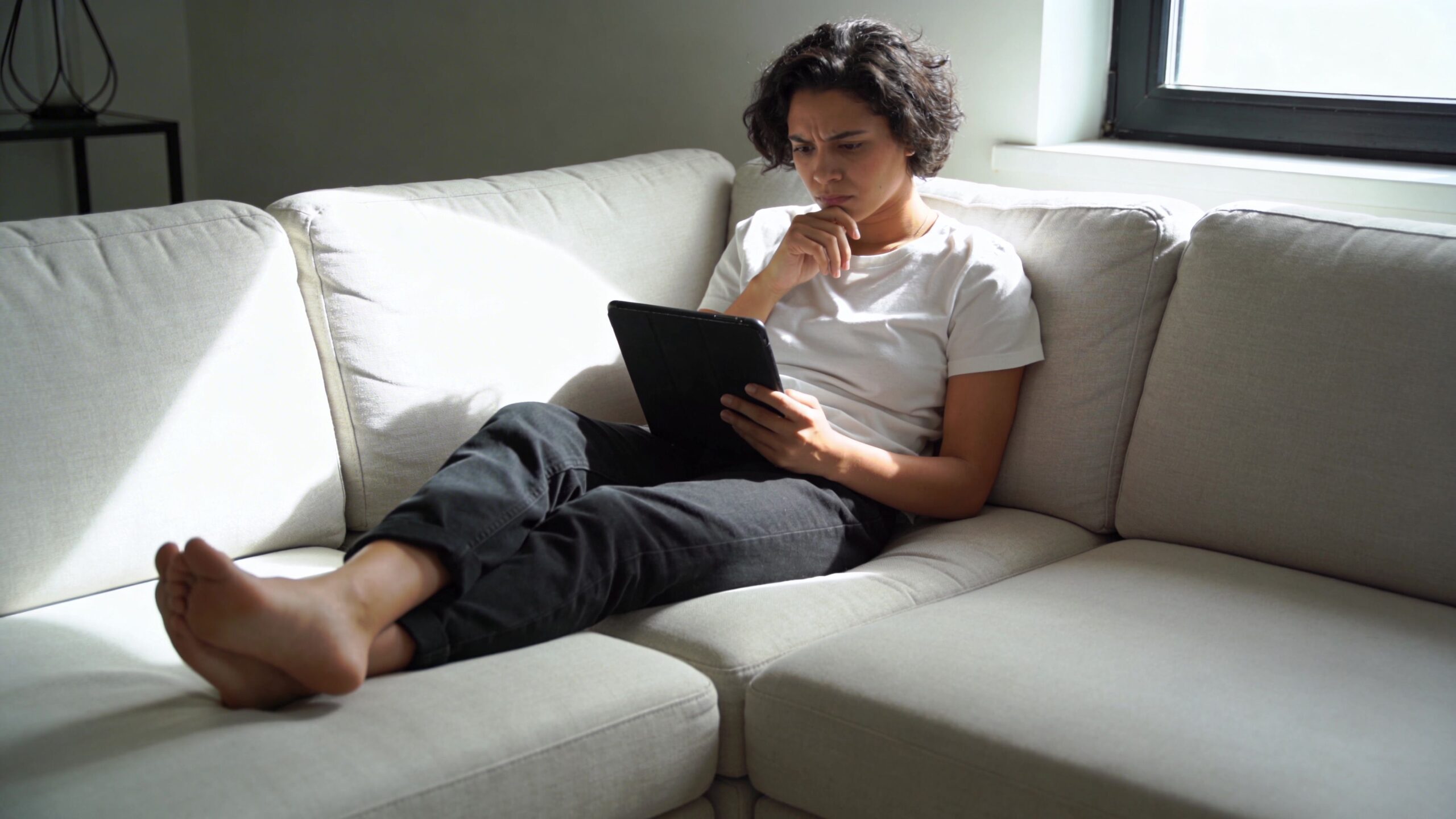 A young woman with curly hair sitting on a comfortable couch while reading information on a tablet.