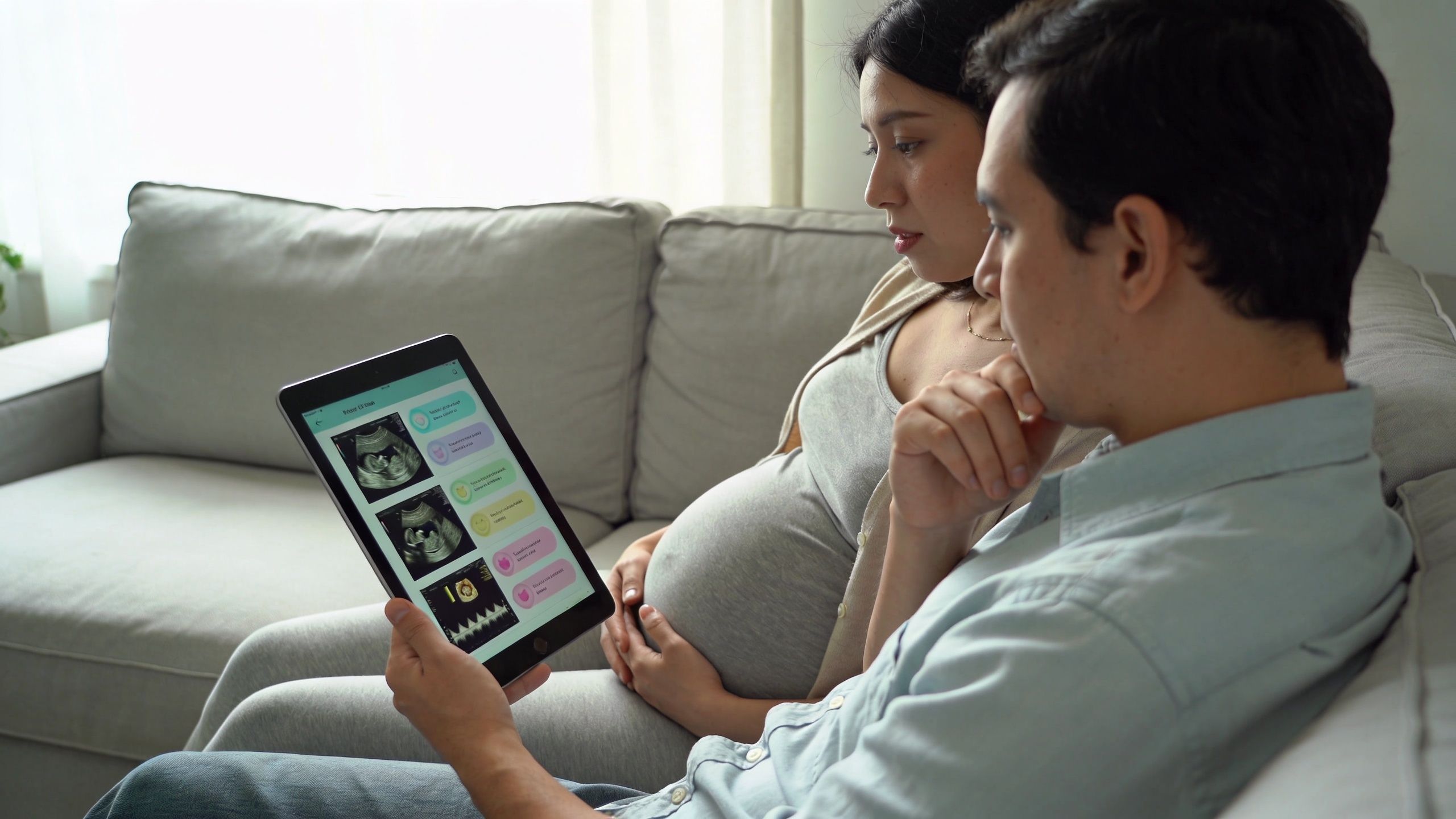 A pregnant woman and her partner sitting on a couch, looking at ultrasound images on a tablet.